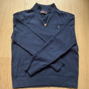 Mörkblå stickad halfzip från Morris - Säljer en mörkblå stickad tröja från Morris med halv dragkedja och hög krage. Tröjan har ribbade muddar och en liten broderad logga på bröstet. Perfekt för kyligare dagar och enkel att matcha.