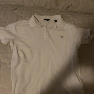 Vit pikétröja från GANT - Säljer en klassisk vit pikétröja från GANT med liten broderad logga på bröstet. Tröjan har krage och korta ärmar, perfekt för en stilren och enkel look. Passar till mycket och är ett måste i garderoben.