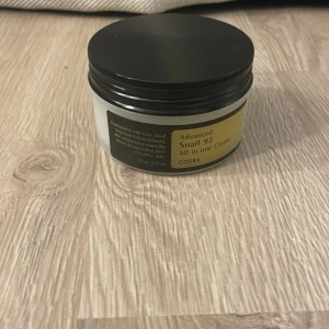 COSRX Advanced Snail 92 All in One Cream - Fuktgivande ansiktskräm med 92% snigelsekret från COSRX. Kommer i en rund, vit plastburk med svart lock. Krämen är vit och har en lätt, gel-liknande konsistens som absorberas snabbt av huden.