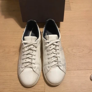 Vita Valentino Garavani sneakers med perforerad V-logga - Säljer ett par vita Valentino Garavani sneakers i skinn med perforerad V-logga på sidorna och svarta detaljer vid hälen. Skorna har rund tå, platt sula och klassisk snörning. Perfekta för dig som gillar stilrena och exklusiva sneakers.
