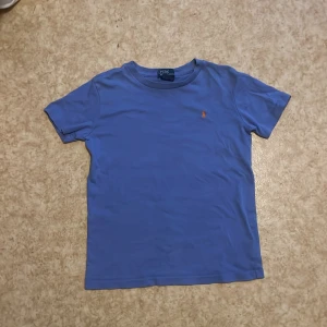 Blå t-shirt från Polo Ralph Lauren - Säljer en blå t-shirt från Polo Ralph Lauren med klassisk orange broderad logga på bröstet. T-shirten har rund hals och korta ärmar, perfekt för en avslappnad stil. Skriv till mig om ni har nån fråga eller nått om tröjan tröjan  eller om storlek för jag vet inte exakt vad storleken på tröjan är!