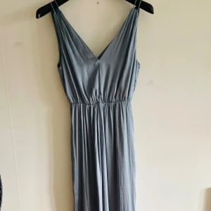 Ljusgrå/blå Långklänning från H&M - Säljer en elegant ljusgrå/blå långklänning från H&M med v-ringning och smickrande midjeresår. Klänningen har ett mjukt, glansigt material som ger en lyxig känsla. Perfekt för speciella tillfällen. aldrig använd! 