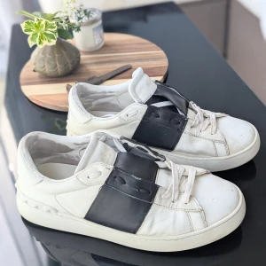 Vita Valentino sneakers  - Säljer ett par vita Valentino sneakers med bred svart läderdetalj över snörningen. Skorna har rund tå, platt sula och klassisk design med diskret logga på plösen. Perfekta för dig som gillar stilrena och exklusiva sneakers. Upplagda på nytt pga oseriös köpare. Så buda inte om du inte står fast vid priset tack 😊