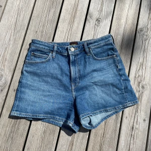 Blå jeansshorts från Lee - Klassiska blå jeansshorts från Lee med fem fickor och hög midja. Shortsen har en normal passform och stängs med knapp och dragkedja. Perfekta för varma dagar och enkla att matcha med olika toppar. Endast provade!