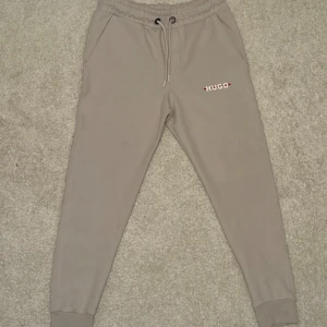 Beige mjukisbyxor från Hugo Boss - Snygga beige mjukisbyxor från Hugo med dragsko i midjan och logga på vänster lår. Byxorna har ribbade muddar vid bensluten och fickor på sidorna samt en bakficka. Perfekta för en avslappnad stil. 