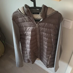 Brun pufferjacka med grå ärmar och huva - Säljer en silver grå cardigan från gran sasso med grå ärmar och huva. Jackan har dragkedja framtill och två fickor med dragkedja. Perfekt för sommarkvällar och är bara en allmänt snygg cardigan