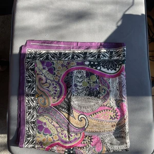 Mönstrad lila och svart halsduk - Säljer en snygg halsduk med paisley- och blommönster i lila, svart, vitt, gult och grönt. Tunt och mjukt tyg med dekorativa detaljer och lila kant. Perfekt för att ge din outfit lite extra färg.