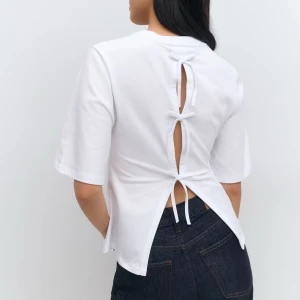 Vit topp  - Säljer en vit topp med kort ärm och unik knytning i ryggen som ger en snygg detalj. Toppen har en rak passform och är tillverkad i mjukt bomullsmaterial. Perfekt för dig som vill ha något stilrent men ändå speciellt.