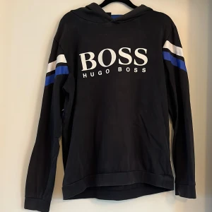 Svart hoodie från Hugo Boss med tryck - Retro hoodie från Hugo Boss, svart  med stort vitt logotryck på bröstet. Tröjan har huva, långa ärmar och vita samt blå ränder på ärmarna. Insidan av huvan är blå. Perfekt för en avslappnad stil. Storlek M.