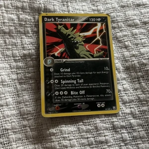 Pokémonkort Dark Tyranitar - Säljer ett samlarkort från Pokémon, Dark Tyranitar med 120 HP. Kortet har svart och gul ram, med en illustration av Tyranitar i mitten. Kortet har attacker som Grind, Spinning Tail och Bite Off. Perfekt för samlare eller Pokémon-fans.
