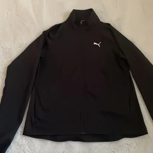 Svart långärmad träningströja från Puma - Säljer en svart långärmad träningströja från Puma med dragkedja och hög krage. Tröjan har en stilren design med Puma-logga på bröstet och är tillverkad i ett stretchigt syntetmaterial.