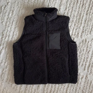 Fleeceväst - Säljer en oanvänd väst med dragkedja och en praktisk bröstficka. Perfekt för kyliga dagar och ger en mysig känsla. Västen har en hög krage för extra värme.