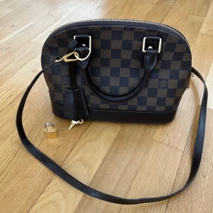 Brun rutig axelväska  - Säljer en klassisk brun axelväska med rutigt mönster och svarta detaljer. Väskan har både handtag och avtagbar axelrem, dragkedja upptill samt medföljande lås och nyckel. Perfekt för dig som gillar tidlös design.