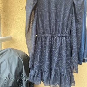 Blå och svart mönstrad klänning med volang - Klänningen har långa, transparenta ärmar och resår i midjan för en snygg passform.