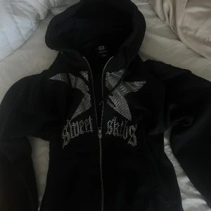 Cool Svart hoodie med strassdetaljer  - Säljer en svart hoodie  med dragkedja och huva. Framsidan har stora silvriga strassdetaljer '. Perfekt statement-plagg för dig som gillar streetstyle.
