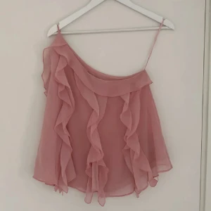 Rosa volang topp med oneshoulder - Säljer en söt rosa topp med volanger och oneshoulder-design. Tunt, luftigt material som ger ett romantiskt intryck. Perfekt för varma dagar eller när du vill känna dig extra fin.