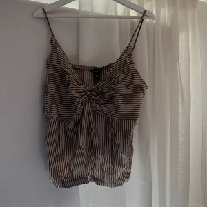 Randig linnetopp från Lindex - Säljer en randig linnetopp från Lindex i beige och svart med smala axelband och knutdetalj framtill. Toppen är ärmlös och har en lätt croppad passform, perfekt för varma dagar. Använd väldigt få gånger, ungefär fem tillfällen. Jag säljer denna för 95 kr + frakt.