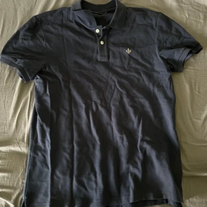 Mörkblå pikétröja från Morris - Säljer en klassisk mörkblå pikétröja från Morris med korta ärmar och krage. Tröjan har knappar framtill och en broderad logga på bröstet. Perfekt för en stilren och avslappnad look. Jag har bara använt denna piken 3 gånger. 