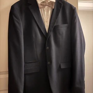 Mörkblå kostym från Selected Homme - Säljer en stilren mörkblå kostym från Selected Homme i slim fit. Kavajen har två knappar, klassiska slag och tre fickor framtill. Byxorna har pressveck och bakficka med knapp. Perfekt för bal eller fest. Tillhörande skjorta och fluga ingår. 
