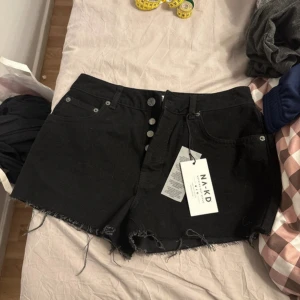 Svarta jeansshorts från NA-KD - Säljer ett par svarta jeansshorts från NA-KD med råa kanter och knappgylf. Klassisk femficksmodell och hög midja. Perfekta för sommaren och lätta att matcha med olika toppar.