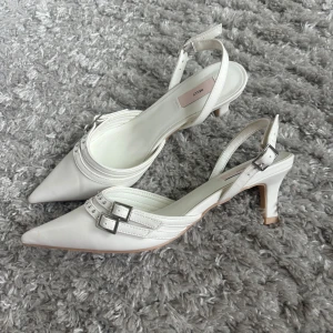 Vita klackskor med spänne från Nelly - Säljer ett par vita klackskor från Nelly med spetsig tå och smala remmar med silverfärgade spännen. Skorna har öppen häl och medelhög klack, perfekta för att lyfta din outfit.
