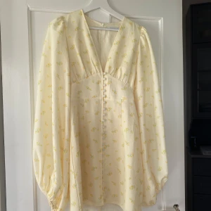 Ljusgul blommig klänning med knappar - Söt ljusgul klänning med små blommor och v-ringning. Klänningen har långa puffärmar och dekorativa knappar framtill. Perfekt för vår och sommar! Använd två gånger💛