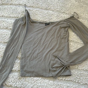 Beige offshoulder topp med lång ärm - Trendig beige topp med offshoulder-design och lång ärm. Tunn och mjuk i materialet, perfekt för att skapa en cool och avslappnad look. Snygg detalj med ena axeln bar. 