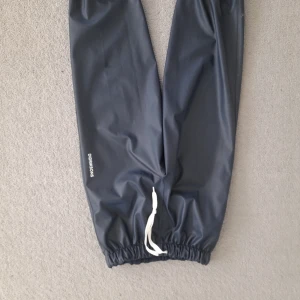 Mörkblå regnbyxor från Didriksons - Mörkblå täckbyxor från Didriksons med resår i midjan och benslut samt vit dragsko framtill. Perfekta för kalla och blöta dagar. Vattentätt material och enkel design.