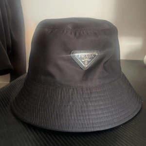 Svart bucket hat från Prada - Snygg svart bucket hat från Prada med klassisk logga framtill. Hatten har en enkel och stilren design i syntetmaterial, perfekt för att lyfta din outfit.