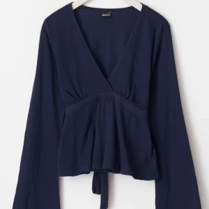 Mörkblå blus med v-ringning och knyt - Snygg mörkblå blus med v-ringning och knyt i midjan. Långa ärmar och lätt utsvängd passform nedtill. Perfekt att styla med jeans eller kjol för en trendig look.