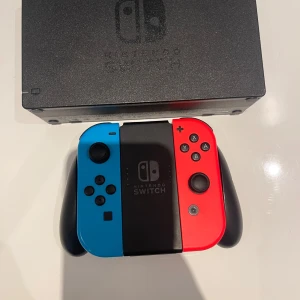 Nintendo Switch med blå och röd Joy-Con pris kan diskuteras. - Nintendo Switch med tillhörande dockningsstation och två Joy-Con kontroller i blått och rött. Konsolen har en svart design och kontrollerna kan användas separat eller tillsammans med handgrepp. Perfekt för gaming och multiplayer.