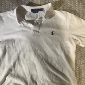 Vit pikétröja från Polo Ralph Lauren - Klassisk vit pikétröja från Polo Ralph Lauren med blå broderad logga på bröstet. Tröjan har korta ärmar, krage och knappar vid halsen. Perfekt för en stilren och tidlös look.