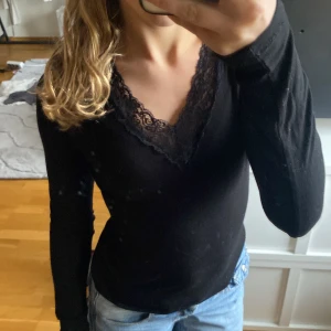 Svart långärmad topp med spets från Gina Tricot - Säljer en svart långärmad topp från Gina Tricot med v-ringning och fina spetsdetaljer längs halsringningen. Perfekt att styla med jeans eller kjol för en snygg look.