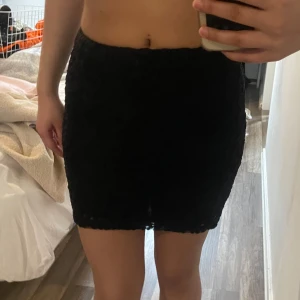 Svart spetskjol - Säljer en svart kortkjol i spets med figurnära passform. Kjolen har en enkel design utan synliga detaljer och är perfekt för dig som gillar en stilren look. Passar till många olika outfits och är lätt att matcha.