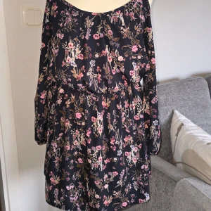 Blommig kortklänning från IJOAH stl L - Supersöt svart kortklänning med rosa och lila blommigt mönster från IJOAH. Klänningen har offshoulder-modell med smala axelband och långa ärmar. Resår i midjan för en bekväm passform. Perfekt för vår och sommar! Klänning är i stl L men kan passa även mindre 