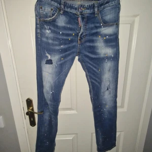 Blå jeansbyxor med färgstänk från Dsquared2 - Säljer ett par blå jeansbyxor från Dsquared2 med slitningar, hål och färgstänk i vitt och gult. Klassisk femficksmodell med rak passform och låg midja. Perfekta för en trendig och avslappnad look.