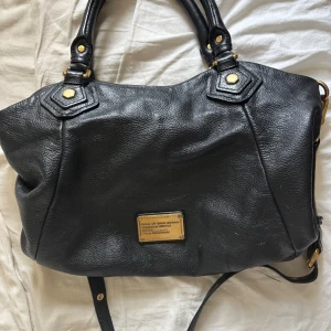 Svart handväska från Marc by Marc Jacobs - Snygg svart handväska i skinn från Marc by Marc Jacobs med guldfärgade metalldetaljer och logoplatta framtill. Väskan har både handtag och axelrem för flexibel användning. Perfekt för dig som vill ha en stilren och rymlig väska. Älskar den men kommer tyvärr inte till användning! 💞