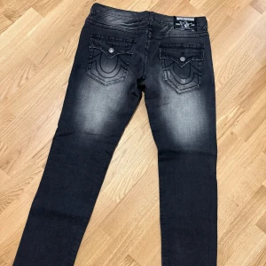 Svarta jeansbyxor från True Religion size 32-36 - Säljer ett par svarta jeansbyxor från True Religion med snygga slitningar och kontrastsömmar. Byxorna har raka ben, klassiska fem fickor och dekorativa bakfickor med lock och knapp. Perfekta för en cool och avslappnad stil.