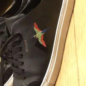 Svarta sneakers från Axel Arigato - Snygga svarta sneakers från Axel Arigato med en broderad fågel i färgglada nyanser på sidan. Skorna har en vit sula och svarta snören, vilket ger en stilren look. Perfekta för en casual stil.