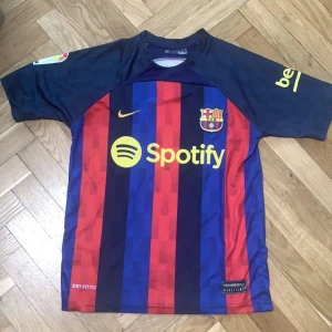 FC Barcelona fotbollströja med Pedri 16 - Säljer en officiell FC Barcelona fotbollströja från Nike med Pedri och nummer 16 på ryggen. Tröjan har ett klassiskt randigt mönster i blått och rött med gula detaljer. Den är kortärmad och har sponsorloggan Spotify på framsidan.