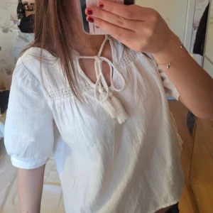 Vit topp med knytband från Vero Moda - Säljer en söt vit topp från Vero Moda med knytband framtill och fina spetsdetaljer vid halsen. Toppen har trekvartsärmar och en luftig passform, perfekt för vår och sommar. 💞