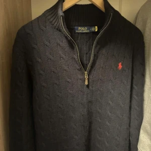 Svart stickad tröja från Ralph Lauren - Säljer en stilren svart stickad tröja från Ralph Lauren med dragkedja och det klassiska röda logotypbroderiet på bröstet. Tröjan har en ribbad krage och långa ärmar, perfekt för kyligare dagar.