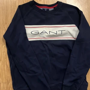 Mörkblå sweatshirt från GANT - Säljer en snygg mörkblå sweatshirt från GANT med ett stilrent tryck på bröstet. Tröjan har långa ärmar och ribbade muddar vid ärmslut och nederkant. Perfekt för en avslappnad stil.