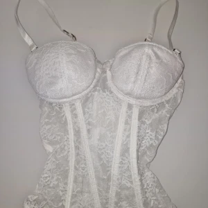 Vit spetskorsett - Vit korsett med spetsdetaljer, justerbara axelband och strumpebandshållare. Korsetten har vadderade kupor och en feminin passform.  