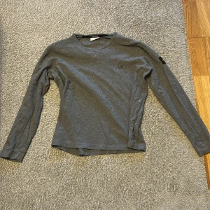 Grå långärmad tröja från Calvin Klein - Säljer en stilren grå långärmad tröja från Calvin Klein. Tröjan har en diskret CK-logga på bröstet och en etikett på ärmen. Perfekt för en avslappnad look.Passar perfekt för personer som har S