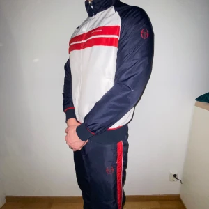 Vit och marinblå träningsjacka från Sergio Tacchini - Snygg tracksuit med prasselmaterial i vitt och marinblått med röda detaljer från Sergio Tacchini. Jackan har en dragkedja framtill och långa ärmar. Perfekt för våren. Den är i nyskick använd 5 gånger då den är förlåten för mig. Den är i storlek L men passar också definitivt någon i M. Bara att höra av sig vid frågor.