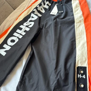 Svarta byxor med tryck från H-4 - Coola svarta byxor från H-4 med stort vitt tryck 'H4 FASHION' längs benet. Byxorna har orange och vita detaljer på sidorna och elastisk midja för extra komfort. Perfekta för en sportig look.