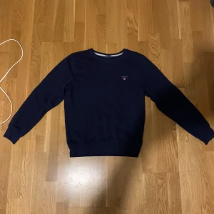 Mörkblå tröja från GANT - Säljer en stilren mörkblå tröja från GANT med broderad logga på bröstet. Tröjan har långa ärmar och ribbade muddar vid ärmslut och nederkant. Perfekt för en avslappnad look.