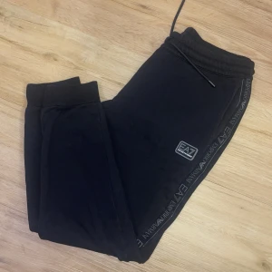 Svarta mjukisbyxor från Emporio Armani - Snygga svarta mjukisbyxor från Emporio Armani med EA7-logga på sidan. Byxorna har en elastisk midja med dragsko och ribbade muddar vid bensluten. Perfekta för en avslappnad stil.