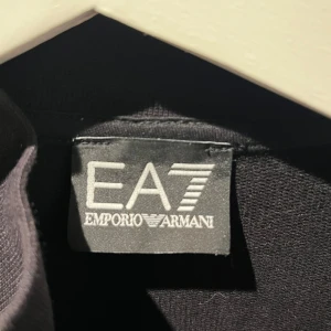 Svart hoodie från Emporio Armani - Säljer en stilren svart hoodie från Emporio Armani med dragkedja framtill. Tröjan har en hög krage och två praktiska fickor. EA7-loggan syns både på bröstet och ryggen. Perfekt för en casual look.